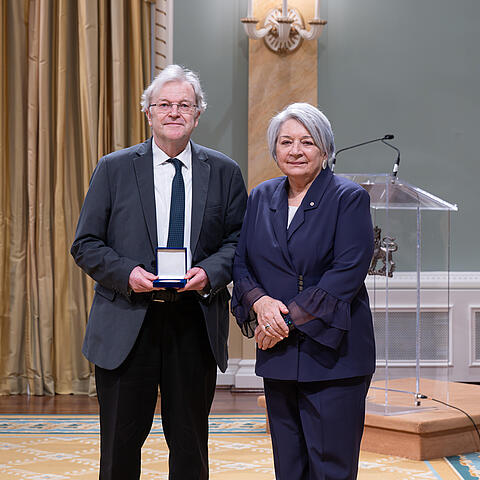Porträtfoto nach einer Preisvergabe: Prof. Dr. Hans-Jürgen Lüsebrink steht links im Anzug und hält ein geöffnetes Etui mit der verliehenen Auszeichnung. Rechts neben ihm steht Mary Simon, Generalgouverneurin von Kanada. Im Hintergrund sind ein Rednerpult und Vorhänge in einem repräsentativen Raum zu sehen.