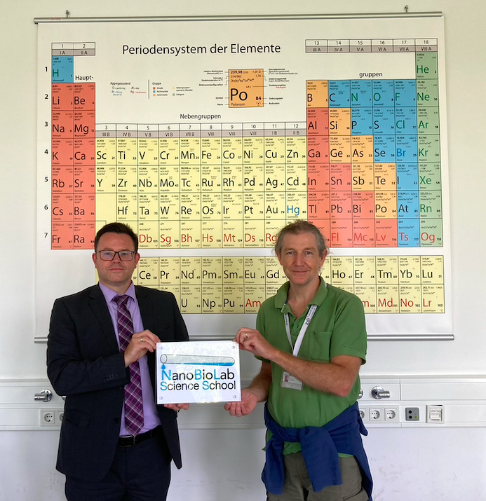 Zwei Personen, die die NanoBioLab Science School Plakette halten, stehen im Chemiesaal vor einem Periodensystem. Links steht der Schulleiter des Marie-Luise-Kaschnitz-Gymnasiums und rechts der Leiter des Arbeitskreises Prof. Kay  