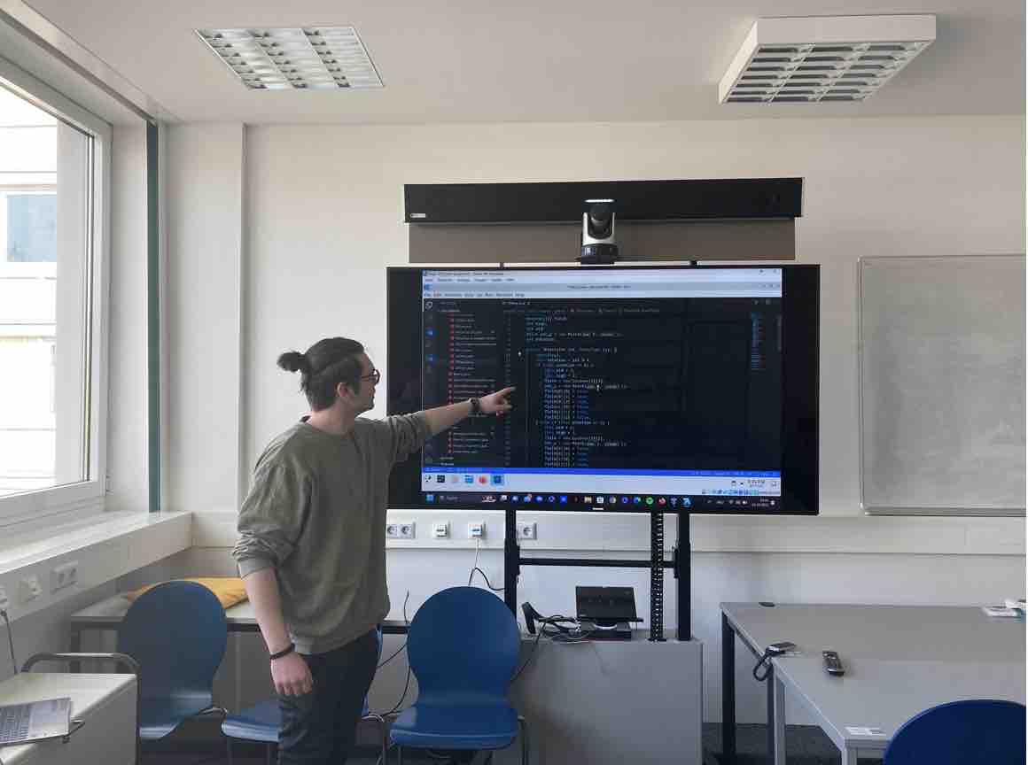 Mensch steht in Seminarraum vor einem Bildschirm und zeigt auf Quellcode