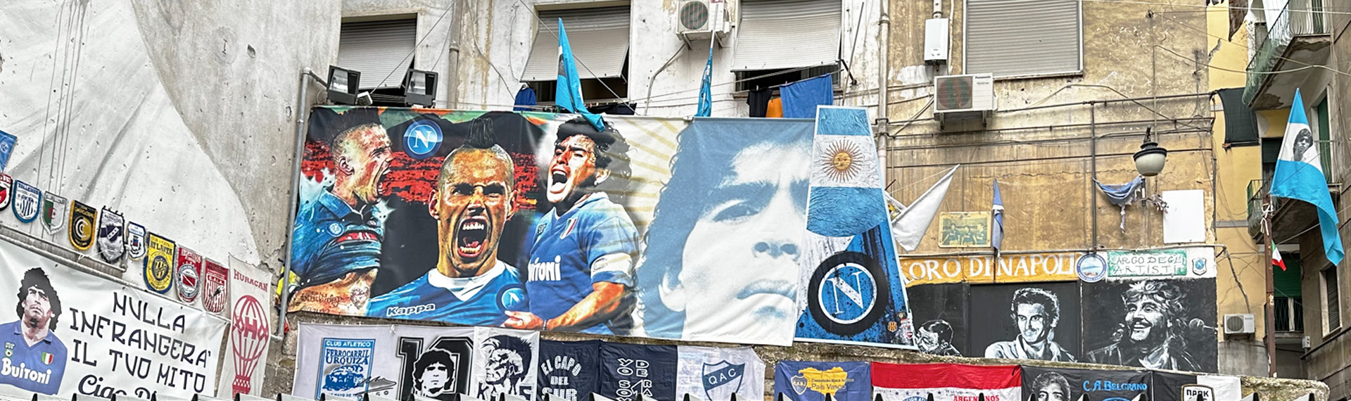 Fotografie einer Stadtmauer geschmückt mit Aufklebern und Postern von fußballmanschaften. Groß in der  MItte ein Poster der Argentinischen Fußballmannschaft.  
