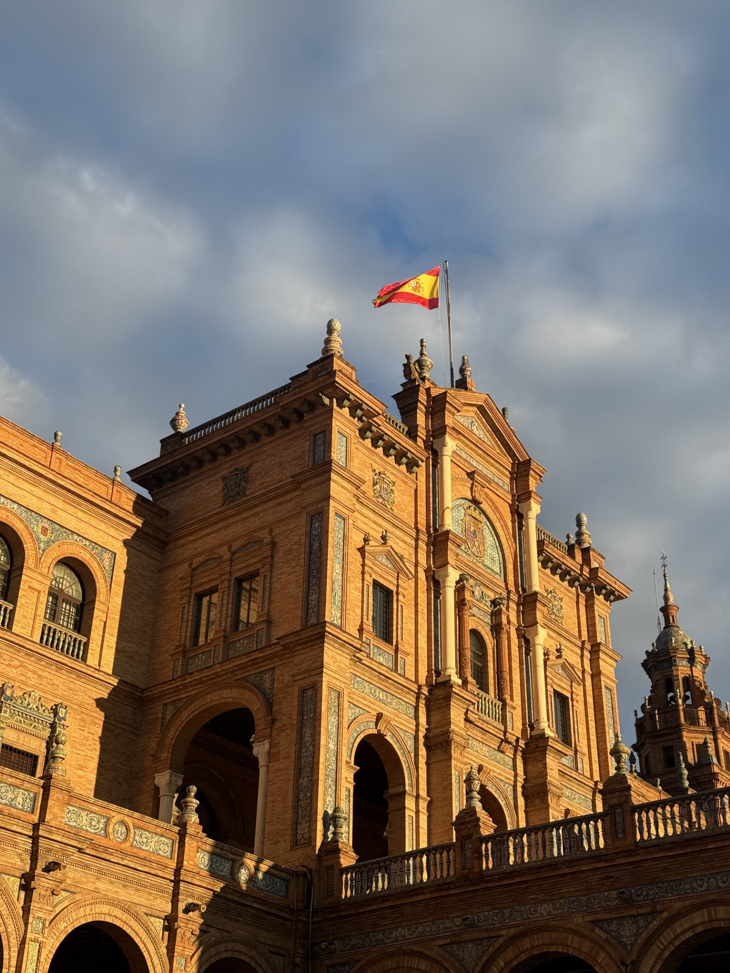 Foto von einem großen Gebäude, angestrahlt von der Sonne, oben drauf eine wehende Spanienflagge.