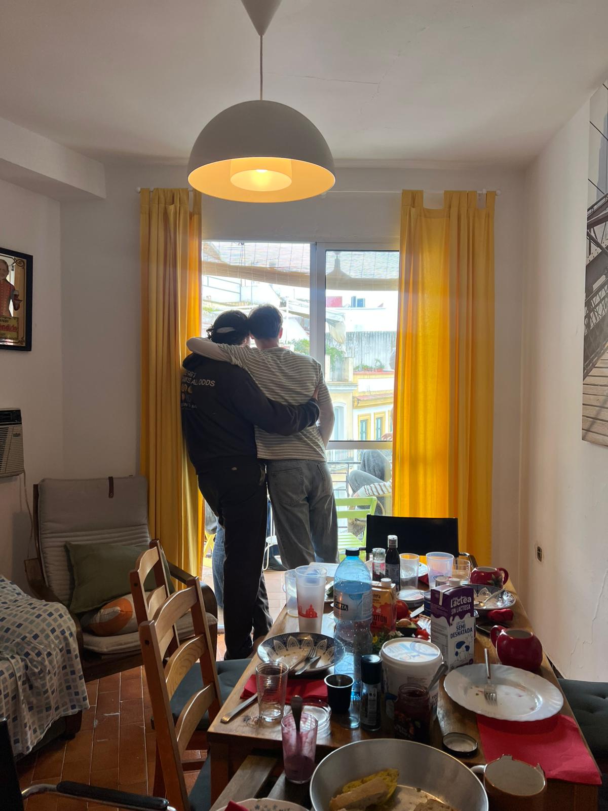 EIn Foto aus einer Wohnung, im Hintergrund ein offenes Fenster mit gelben Vorhängen an der seite, davor zwei Personen, die sich umarmen. Im Vordergrunf ein gedeckter Tisch