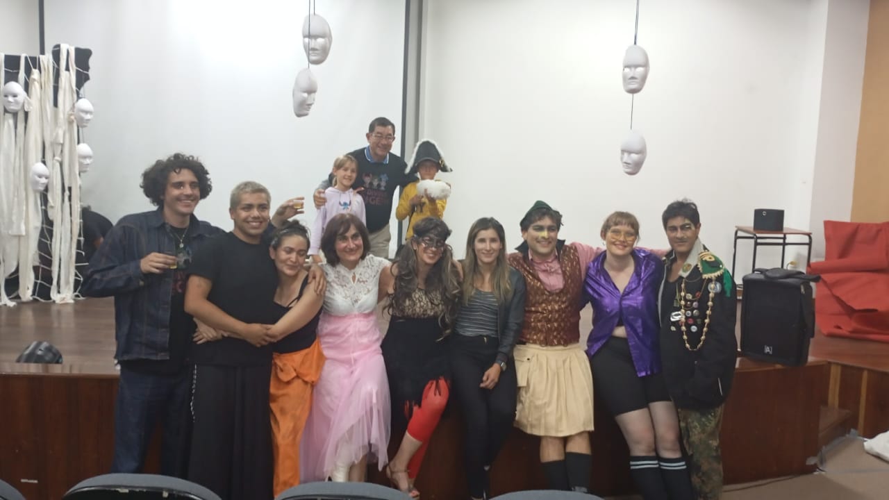 Gruppenfoto von 11 Personen nebeneinander vor einer Bühne. Hintergrund: weiße Wand, weiße Masken, die an Stricken von der Decke hängen.