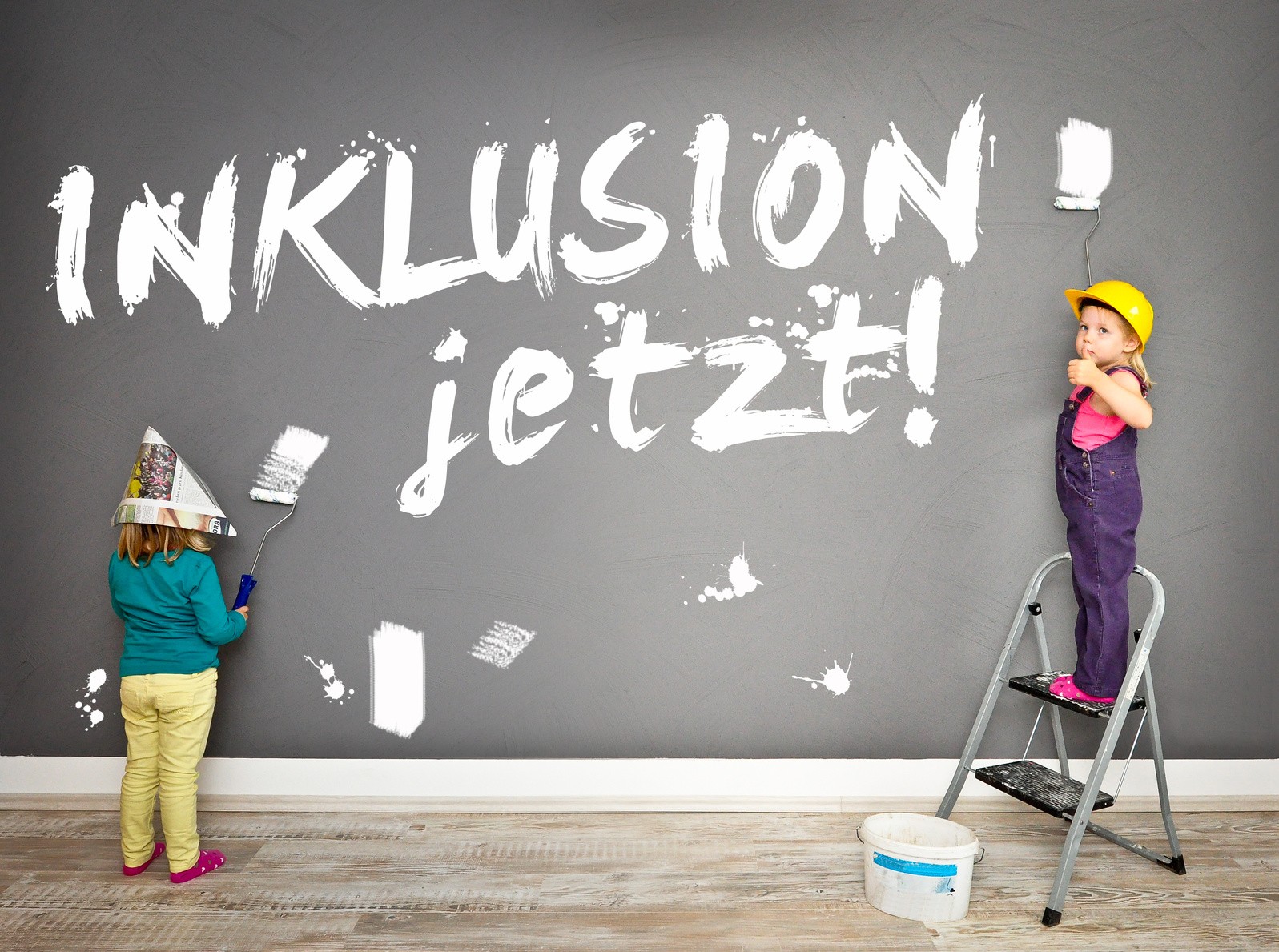 Zwei Kinder in Malerkleidern haben mit weißer Farbe "INKLUSION jetzt!" auf eine dunkle Wand geschrieben.
