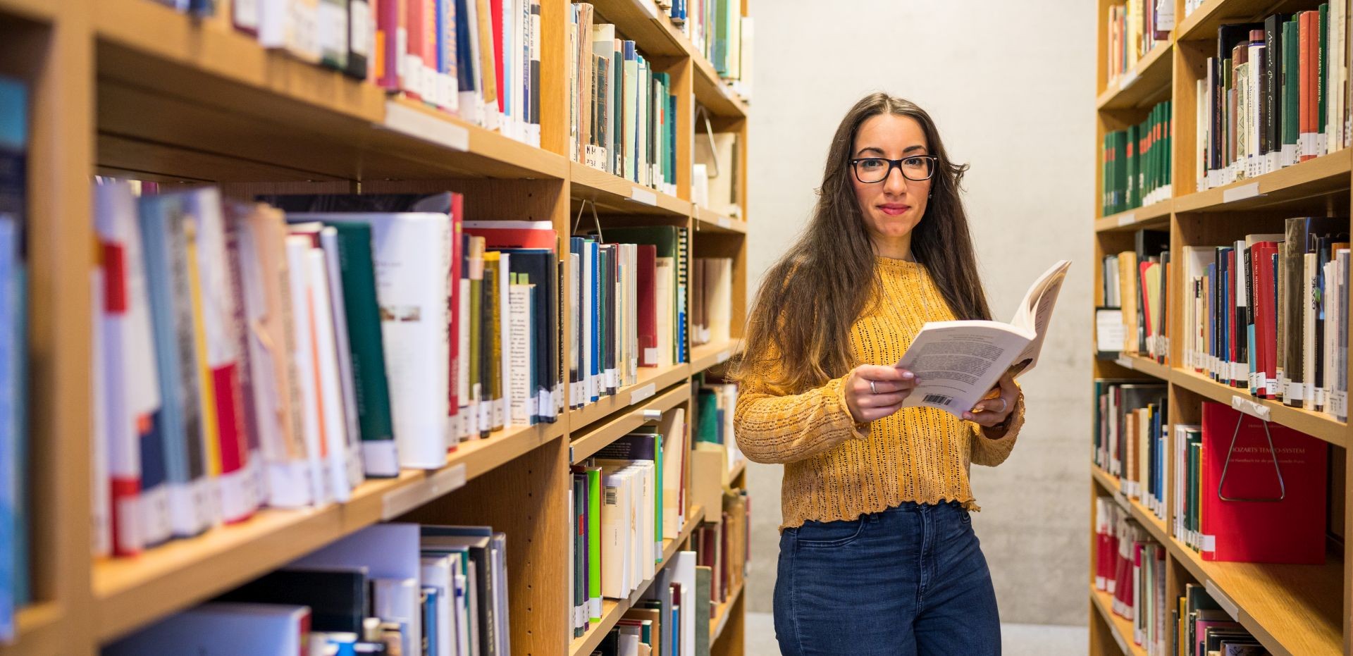 Studentin steht mit einem Buch zwischen zwei Bücherregalen 