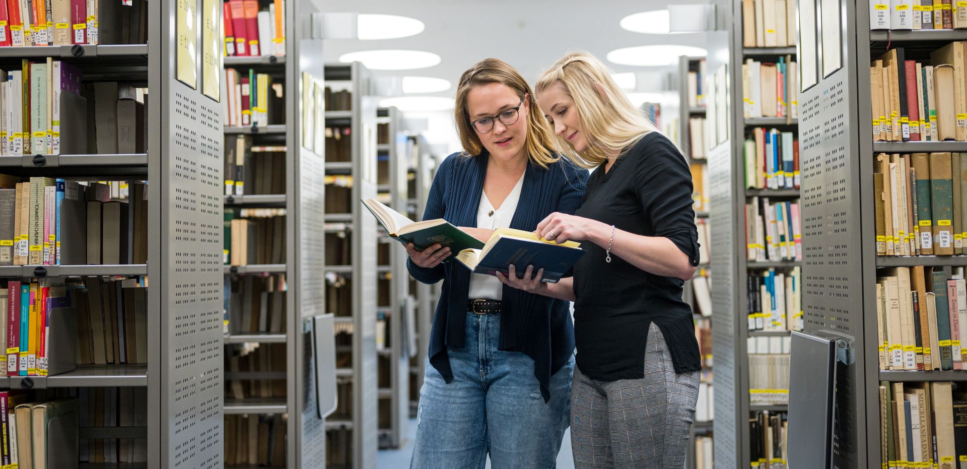 Zwei Studentinnen beraten sich zwischen Bücherregalen in einer Bibliothek