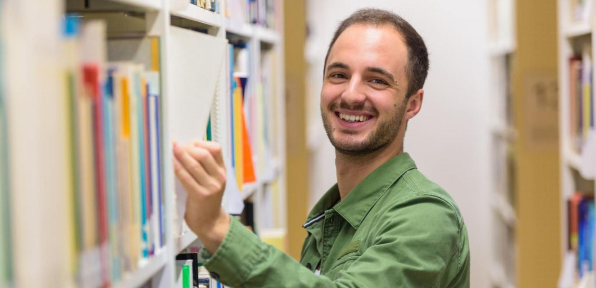 Student steht lächelnd zwischen zwei Bücherregalen