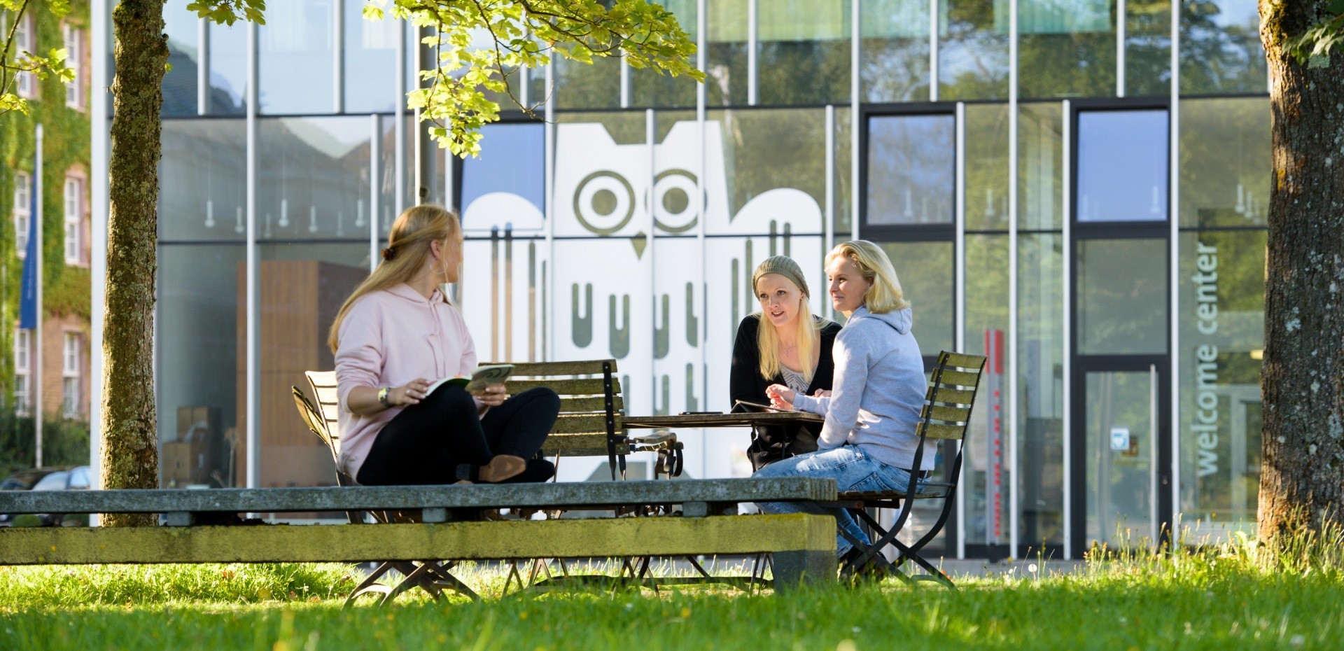 Drei Studentinnen sitzen am Tisch vor einem Glasbau mit großer Campuseule auf den Fenstern