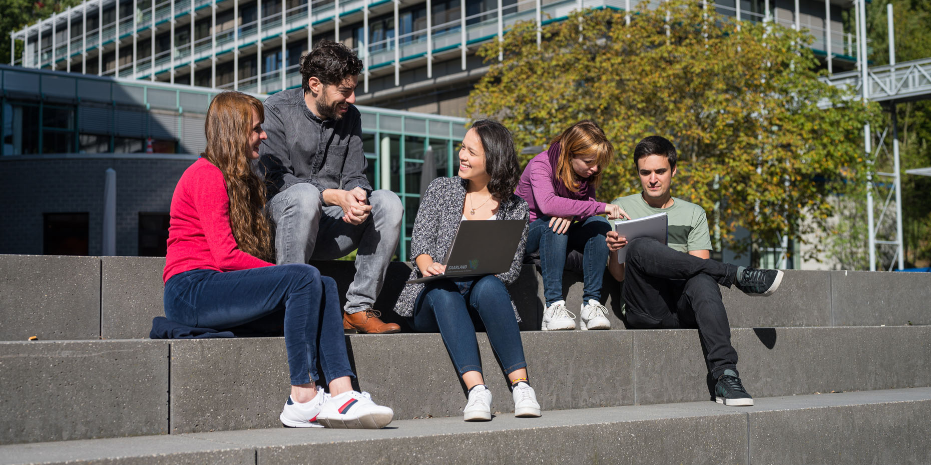 Studierende sitzen auf einer Treppe, unterhalten sich oder schauen in Laptops