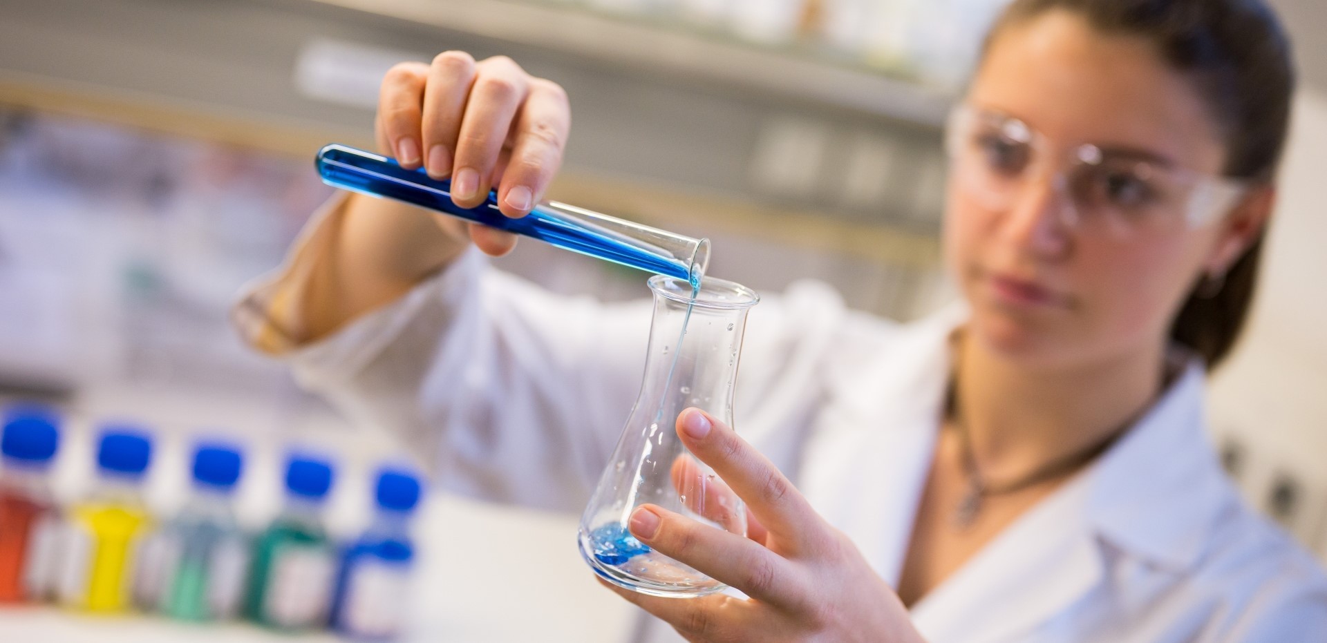 Studentin kippt eine blaue Flüssigkeit aus einem Reagenzglas in einen Glasbehälter