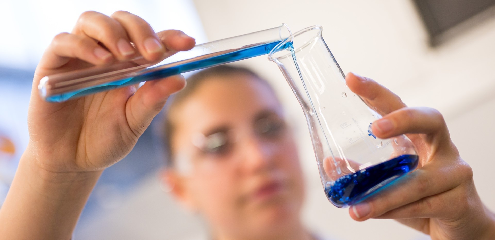 Studentin kippt eine blaue Flüssigkeit aus einem Reagenzglas in einen Glasbehälter