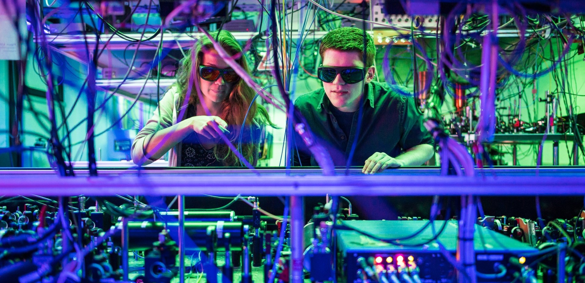 Zwei Studierende stehen mit Schutzbrillen hinter vielen Kabeln in einem Maschinenraum
