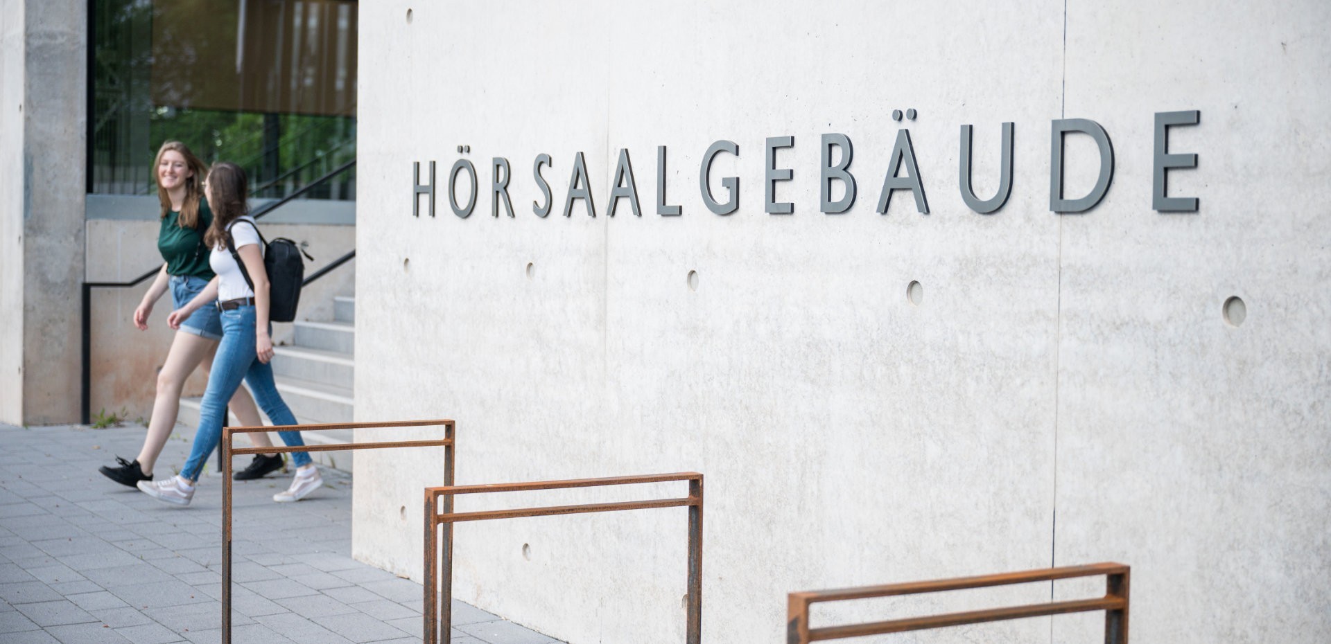 Zwei Studentinnen laufen aus einem Bau mit der Aufschrift "Hörsaalgebäude"