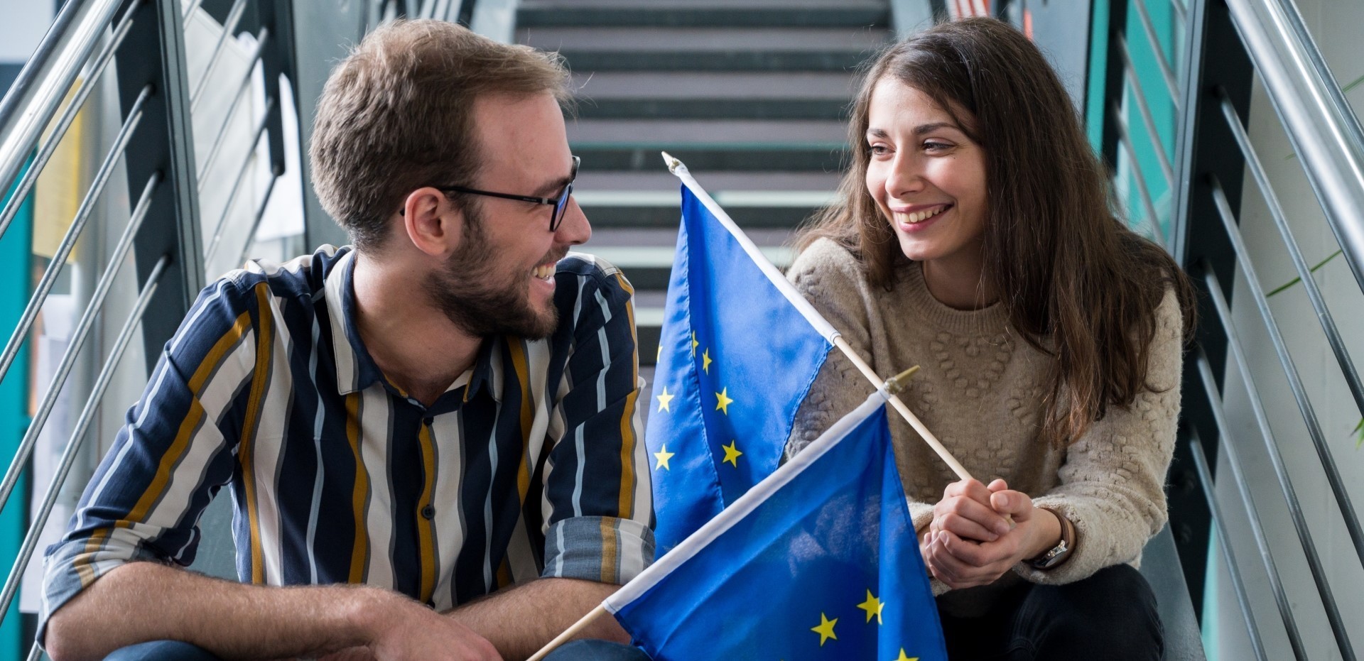 Zwei Studierende sitzen auf einer Treppe und unterhalten sich mit je einer kleiner Europafahne in der Hand