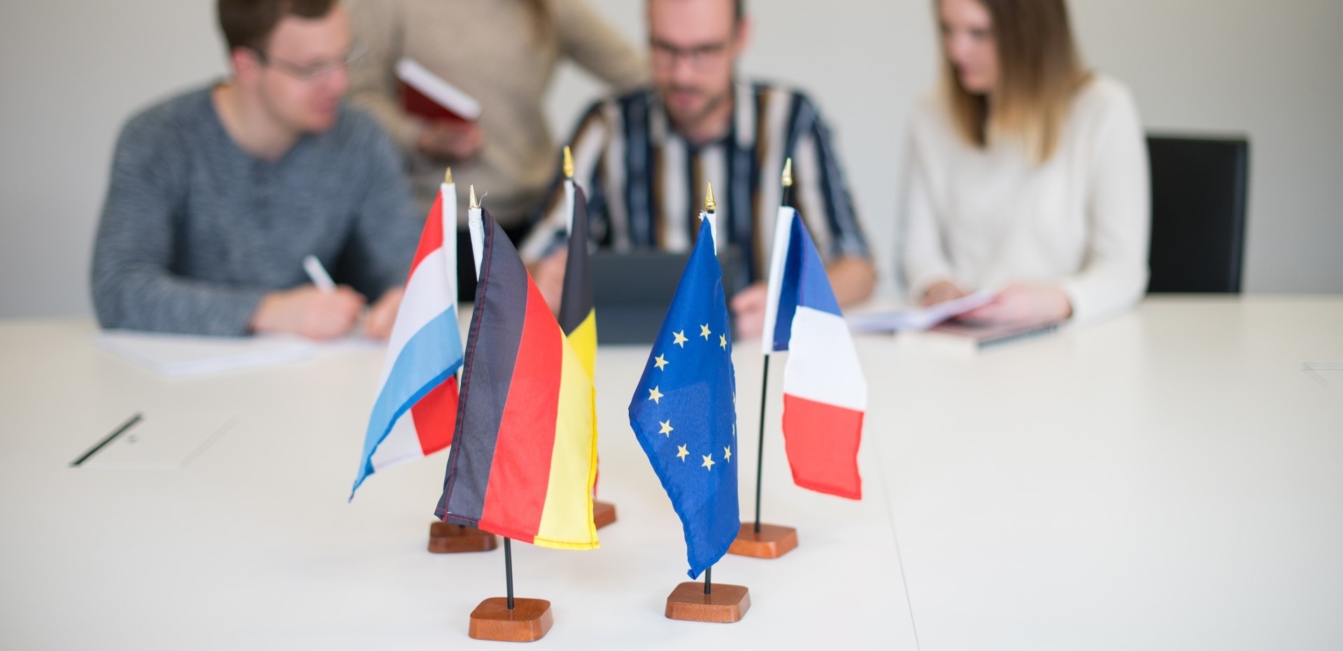 viele kleine Länder-Flaggen auf einem Schreibtisch, dahinter Studenten in einer Besprechung