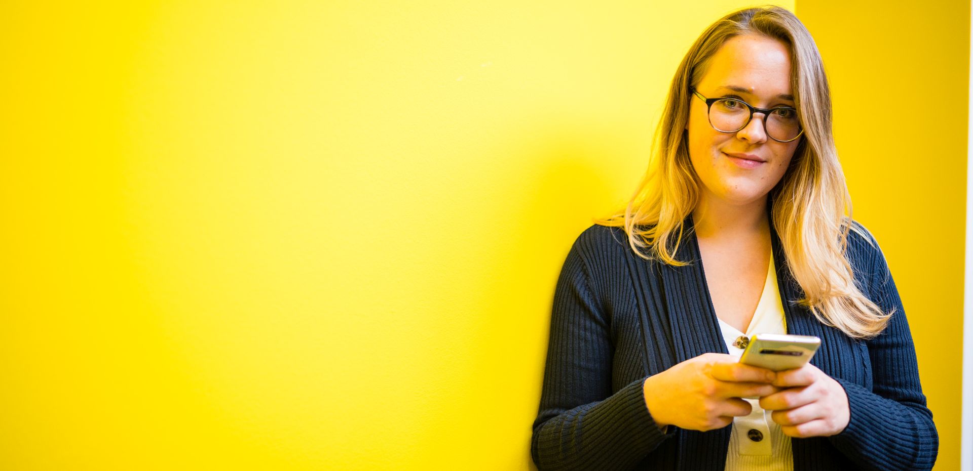 Studentin steht mit Handy vor einer gelben Wand