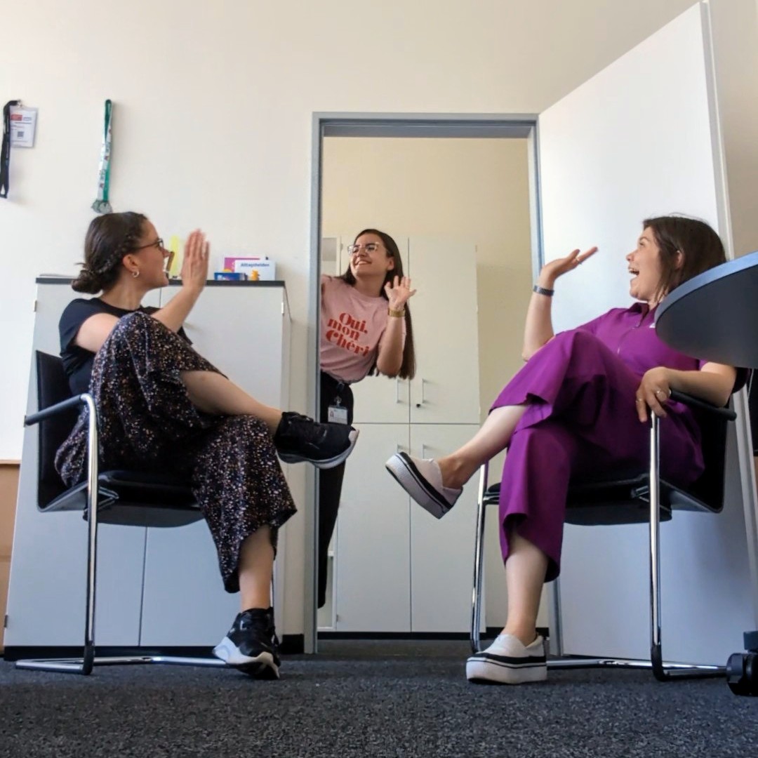 Szene im Büro. Zwei Kolleginnen sitzen im Vordergrund auf Stühlen, eine Kollegin beugt sich durch die Tür hinter ihnen und winkt beiden zu.
