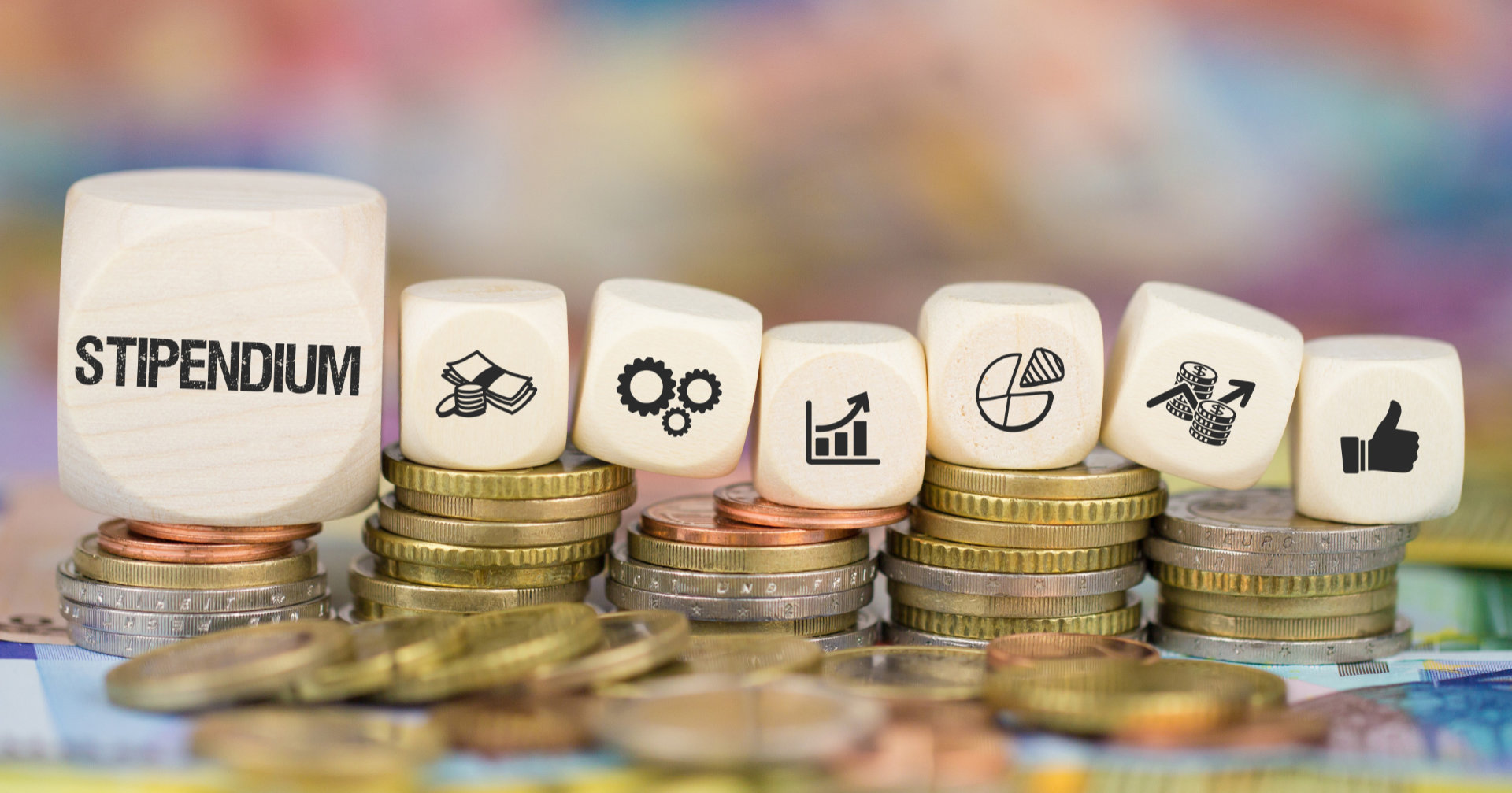 Verschiedene Würfel mit Geld-Symbolen, der größte hat den Aufdruck Stipendium, auf Geldmünzen angeordnet