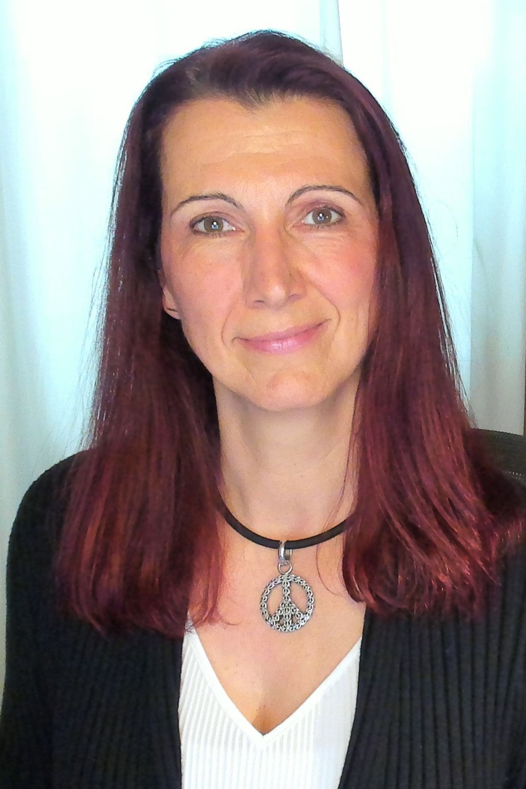 Portrait einer lächelnden Frau mit rotgefärbten Haaren. Sie trägt ein weißes Oberteil und eine schwarze Strickjacke.