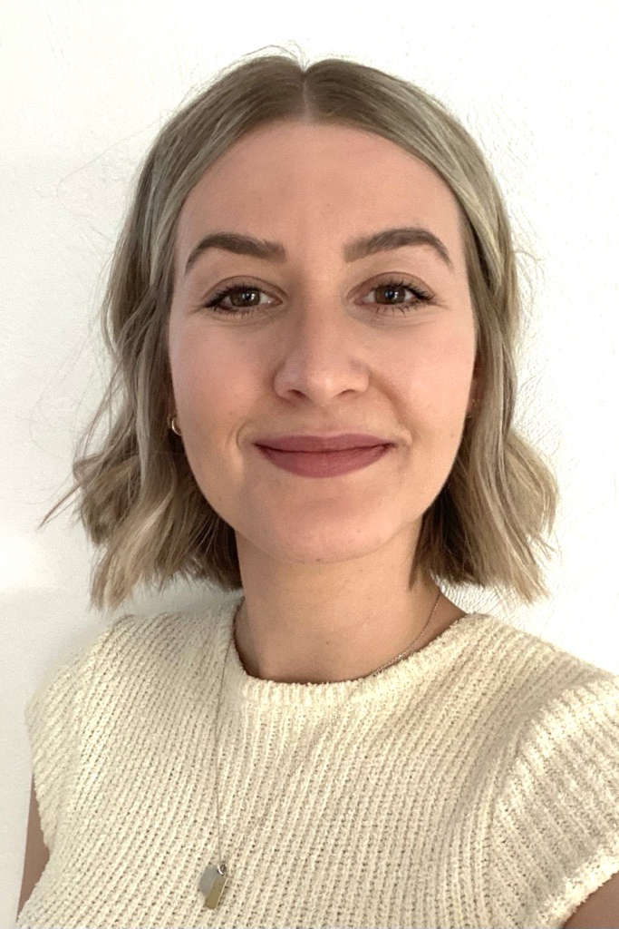 Portrait einer lächelnden Frau mit blondgefärbten Haaren. Sie trägt ein weißes Oberteil.